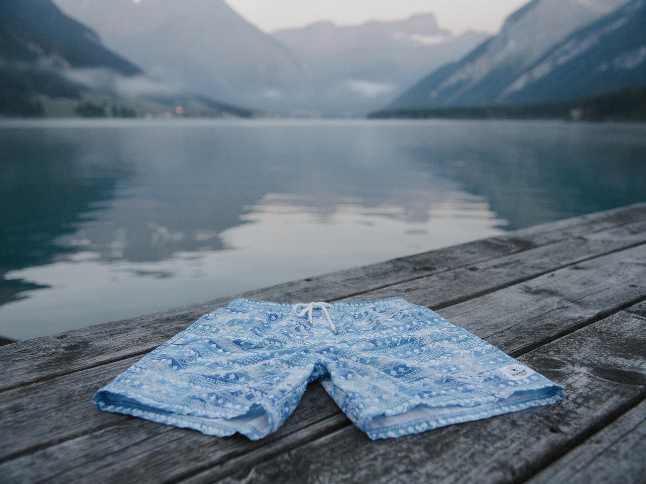 Badehose Greek Azure flach auf einem Holzsteg am Schweizer Alpensee mit Bergen und Nebel im Hintergrund