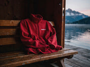Badmeister Fleecejacke rot drapiert auf einer Holzbank in einem Schweizer Bootshaus mit Blick auf Alpensee und Berge in der Dämmerung