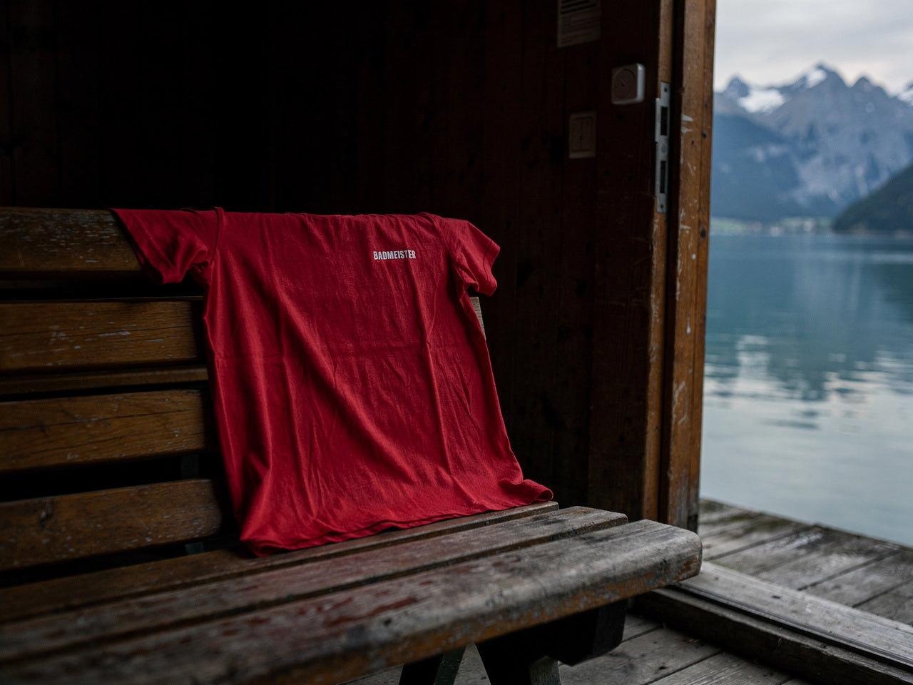 Badmeister T-Shirt rot über einer Holzbank in einem Schweizer Bootshaus mit Blick auf den Alpensee und Berge
